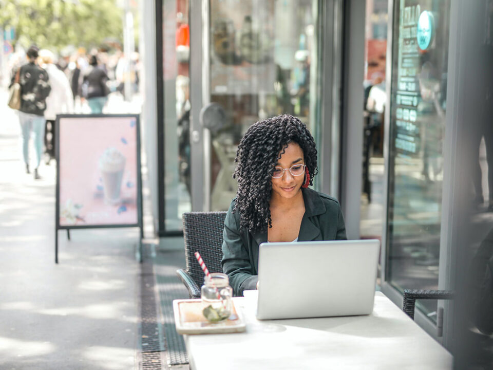 ethnic young woman using laptop to start an online business while having tasty beverage in modern street cafe